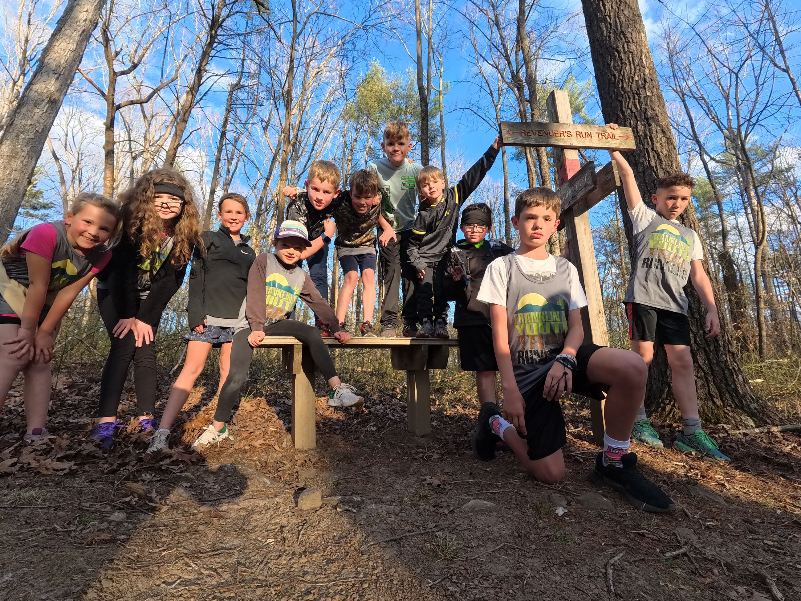 a group of trail runners pose for a photo on a bench and near a trail sign on a trail in the woods